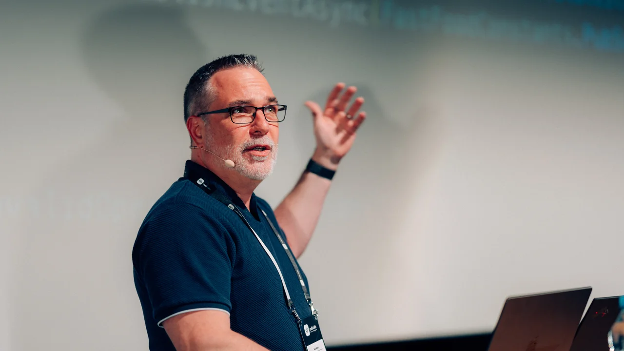 Marc Müller spricht mit Headset vor einer technischen Projektion und einem geöffneten Laptop auf einer Konferenz.